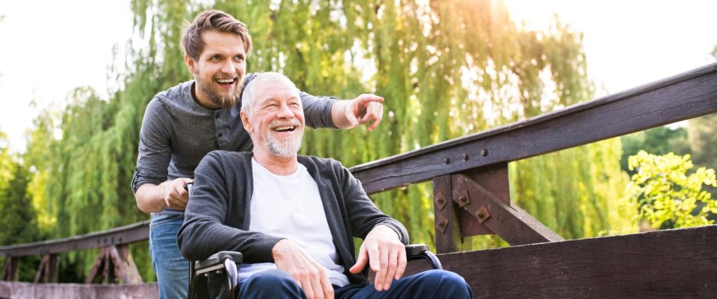 A younger man pushes an older man in a wheelchair on a wooden bridge, both smiling and looking into the distance, with trees in the background.