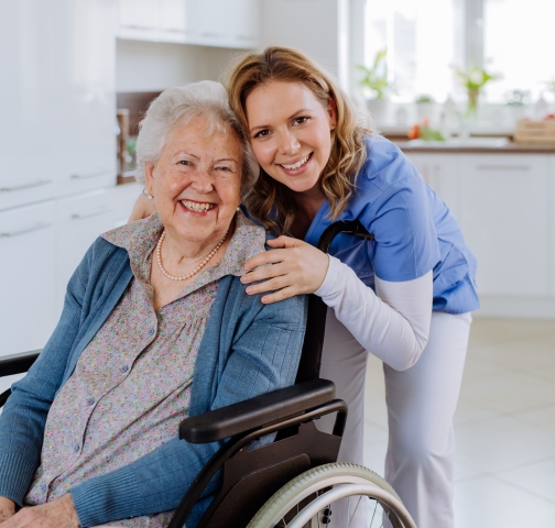 A smiling elderly woman in a wheelchair is embraced by a young caregiver in a blue uniform in a bright indoor setting.