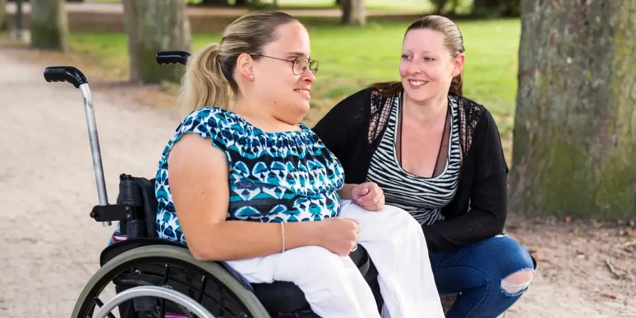 Two women are outdoors; one is sitting in a wheelchair, and the other is kneeling beside her. They are smiling and appear to be in a park with trees in the background.