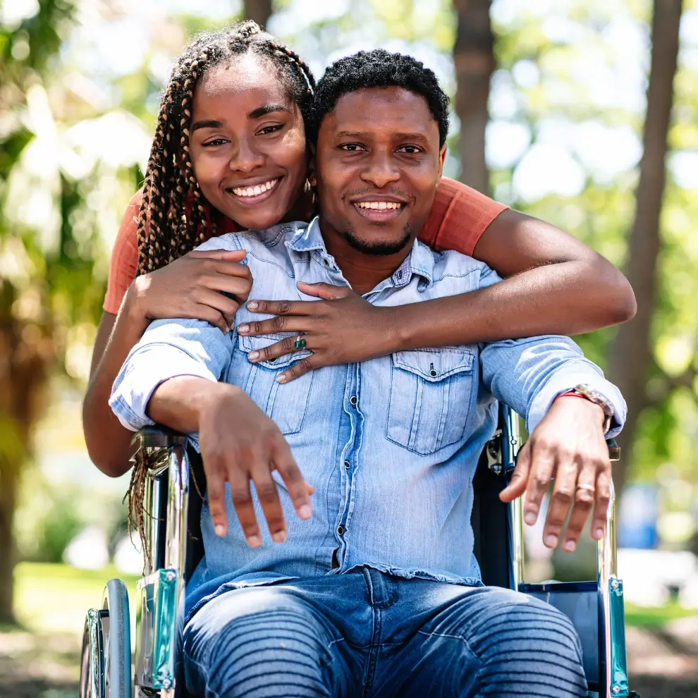 A smiling woman hugs a man from behind. The man is sitting in a wheelchair outdoors on a sunny day.