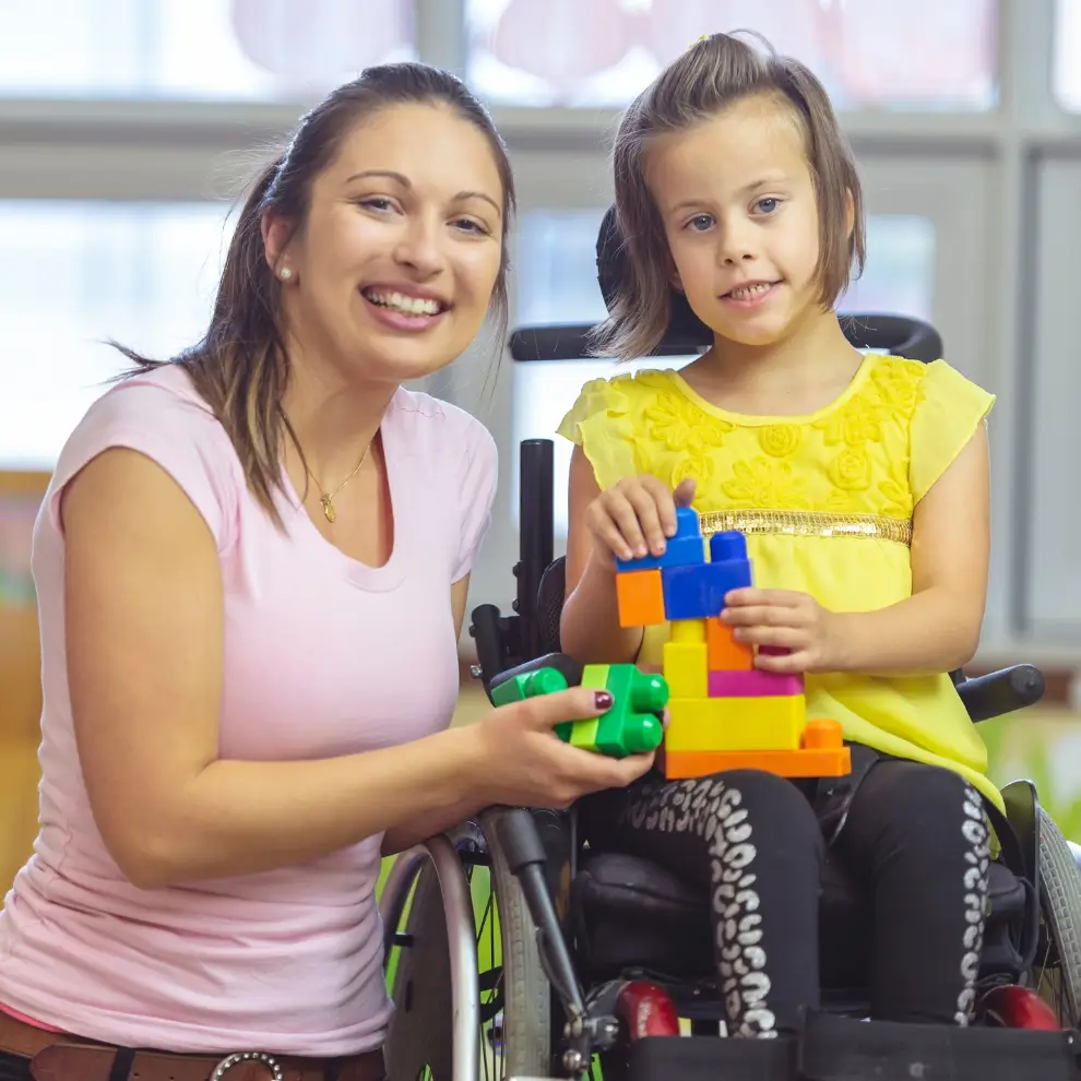 A woman sits beside a young girl in a wheelchair. They are smiling and playing with colorful plastic blocks indoors.