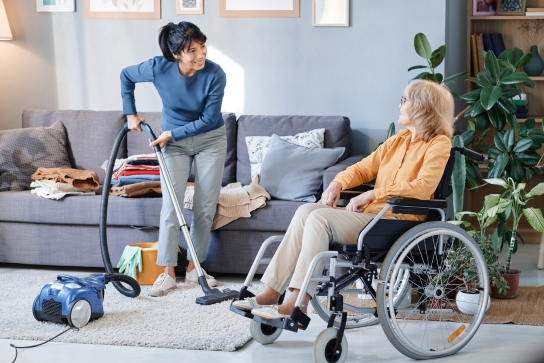 Person vacuuming while talking to a person sitting in a wheelchair in a living room with a sofa and plants.