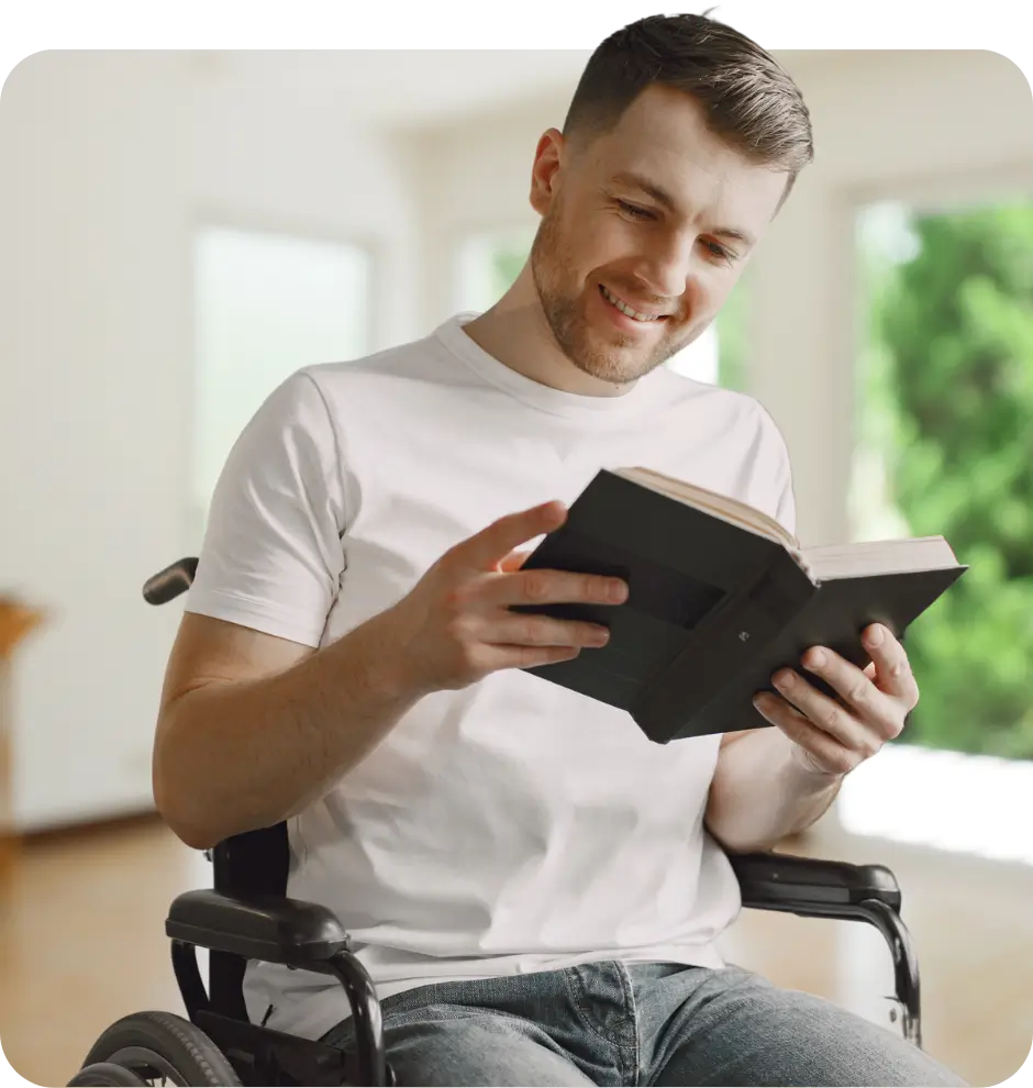 Man sitting in a wheelchair, wearing a white t-shirt and jeans, smiling while reading a book in a well-lit room.